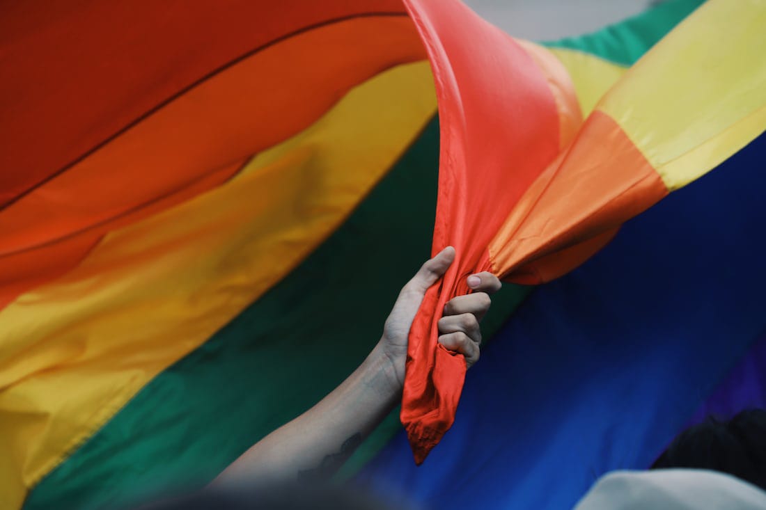 A hand grasping a rainbow pride flag against a blue sky.